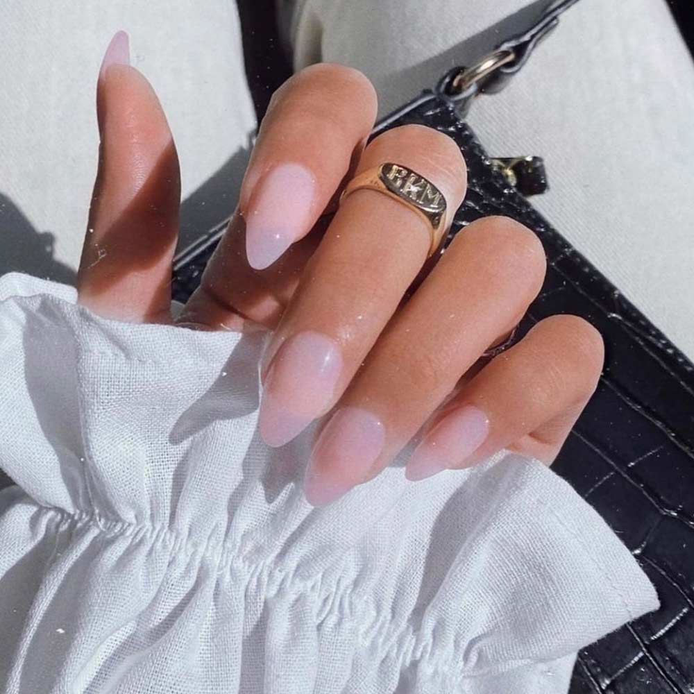 A hand with long, almond-shaped nails in light pink—Cloud 9 by glamnetic—rests on a textured black bag. A gold ring and ruffled white sleeve complete the look, highlighting a chic and elegant manicure.